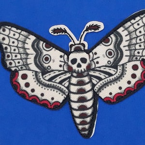 May include: A white and black embroidered moth with a skull design and red accents. The moth is on a blue background.