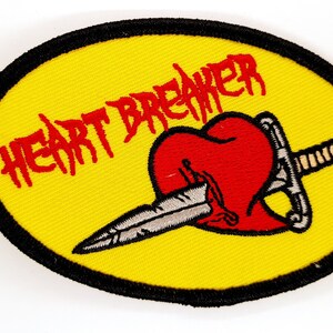 May include: A yellow embroidered patch with a black border. The patch features a red heart with a silver dagger through it. The text "HEARTBREAKER" is written in red above the heart.