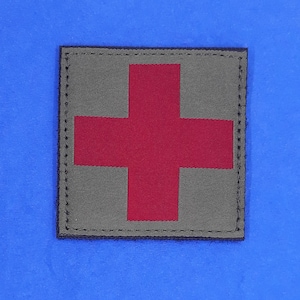 May include: A gray fabric patch with a red cross in the center. The patch has a black stitched border.