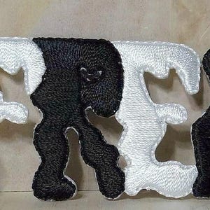 May include: Embroidered patch spelling out the word "FREAK" in a stylized font. The letters alternate between white and black. A U.S. quarter coin is next to the patch for size comparison. The patch is on a light brown surface.