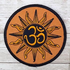 May include: A round, orange embroidered patch with a black outline. The patch features a stylized sun with a black and yellow Om symbol in the center.