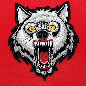 May include: Embroidered patch featuring a snarling wolf's head. The wolf has white fur, black accents, and bright yellow eyes. The mouth is open, revealing sharp white teeth and a red interior. The patch is set against a red background.