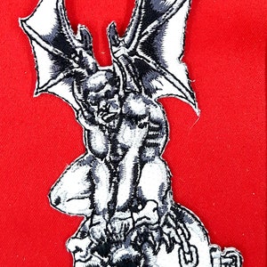 May include: A black and white embroidered patch of a gargoyle with wings sitting on a skull with a chain. The gargoyle is holding the skull with its hands.