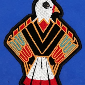 May include: Embroidered patch featuring a stylized eagle design. The patch has a black background with a white eagle head, red beak, and orange and teal accents. The eagle's chest has a white and red pattern.