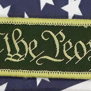 May include: A green fabric patch with yellow stitching around the edges. The patch has the words "We the People" embroidered in a yellow script font.