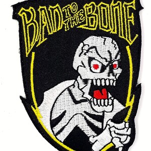 May include: Black and yellow embroidered patch with a white skeleton holding a lightning bolt. The text "Bad to the Bone" is written in yellow above the skeleton.