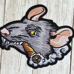 May include: Embroidered patch featuring a gray rat with a cigar in its mouth. The rat has a detailed design with a black outline, pink nose and ears, and a yellow eye. Smoke and a rose are also included in the design.