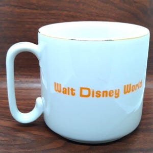 K&ouml;nnte beinhalten: Wei&szlig;e Keramiktasse mit goldfarbenem Rand und dem Schriftzug "Walt Disney World" in orangefarbener Schrift.