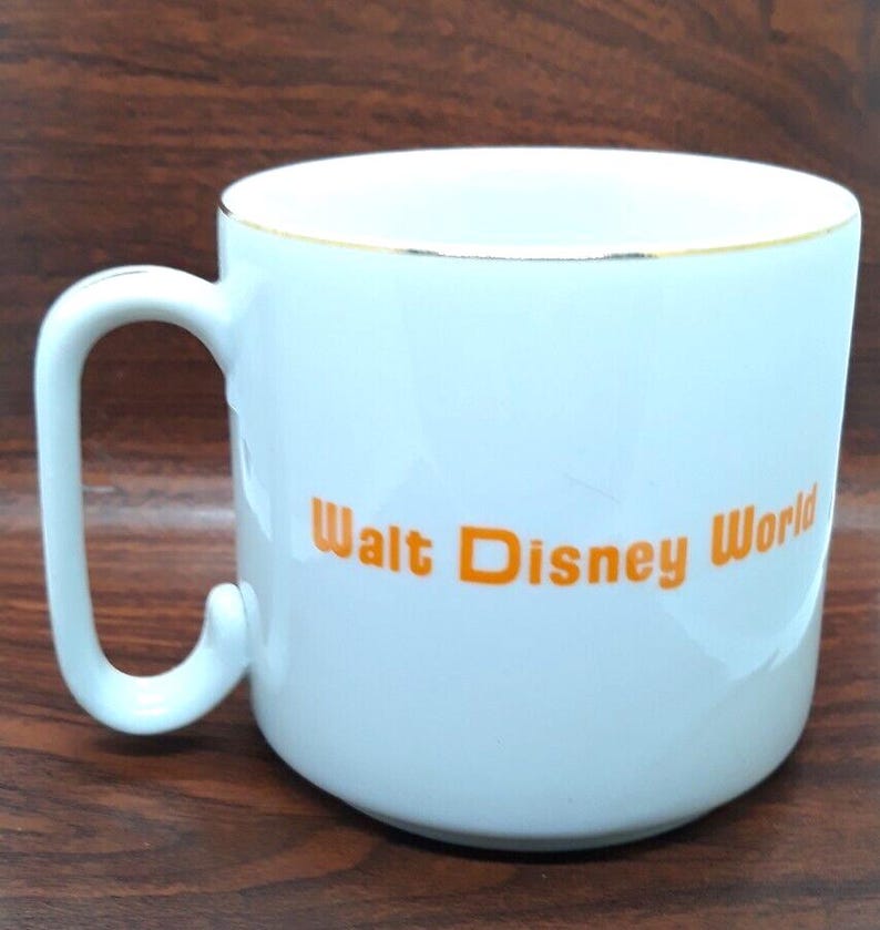 K&ouml;nnte beinhalten: Wei&szlig;e Keramiktasse mit goldfarbenem Rand und dem Schriftzug "Walt Disney World" in orangefarbener Schrift.