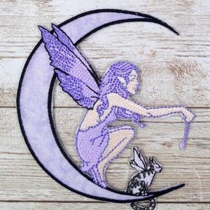 May include: An embroidered patch featuring a light purple crescent moon with a black border. A fairy with purple hair and wings sits on the moon, holding a thin wand. A small winged cat with black and white markings sits below the fairy.