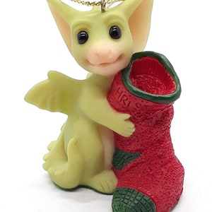 May include: A green dragon figurine holding a red Christmas stocking with green trim. The stocking has the word "CHRIST" printed on it.
