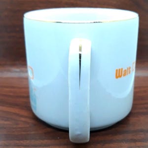 K&ouml;nnte beinhalten: Wei&szlig;e Keramiktasse mit goldfarbenem Rand und Henkel. Die Tasse hat ein orange-blaues Design mit dem Text "Walt Disney World" an der Seite.