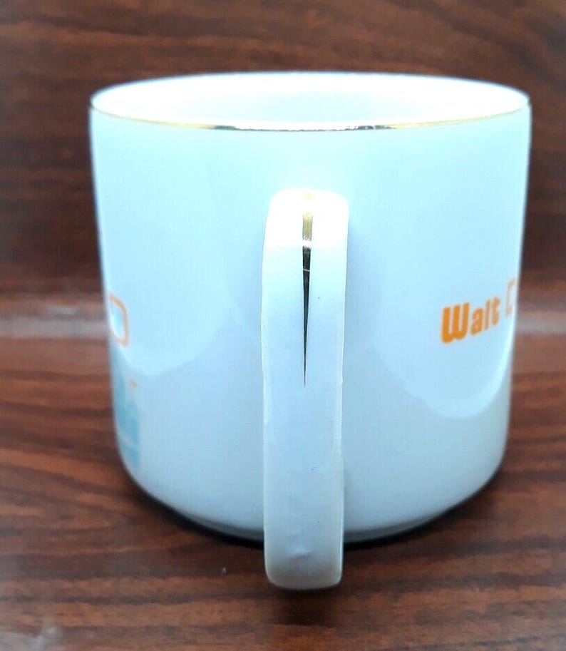 K&ouml;nnte beinhalten: Wei&szlig;e Keramiktasse mit goldfarbenem Rand und Henkel. Die Tasse hat ein orange-blaues Design mit dem Text "Walt Disney World" an der Seite.