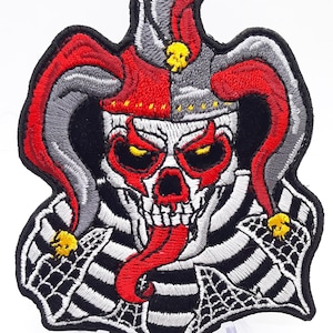 May include: Embroidered patch featuring a skull wearing a jester's hat with red and gray bells. The skull has a red tongue sticking out and is surrounded by a black and white striped pattern with a spider web design.