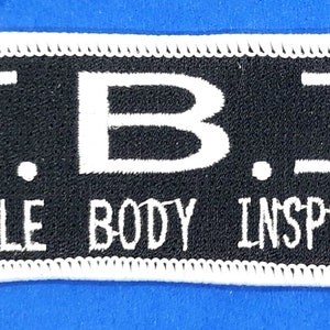 May include: A black and white embroidered patch with the letters "F.B.I." in white and the words "FEMALE BODY INSPECTOR" in white below.