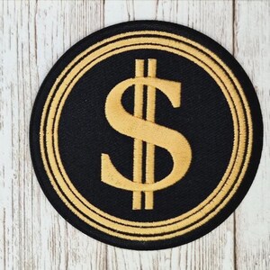 May include: Embroidered patch featuring a gold dollar sign ($) on a black circular background, framed by two gold concentric circles. The patch is likely for clothing or accessories.