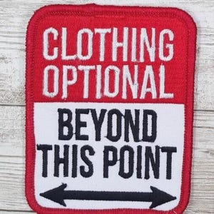 May include: A red and white embroidered patch with the words "CLOTHING OPTIONAL" above "BEYOND THIS POINT" and a black arrow. The patch has a red border and is rectangular in shape.
