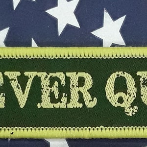 May include: A green and yellow embroidered patch with the text "NEVER QUIT." The patch is on a blue and white American flag background.