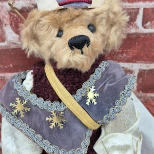 May include: A plush teddy bear wearing a purple velvet outfit with gold snowflake accents and a matching hat. The bear has a black nose and is set against a red brick background. The outfit includes a burgundy knitted vest and gold trim.