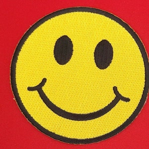 May include: A yellow smiley face patch with a black outline and black eyes. The patch is made of embroidered fabric.