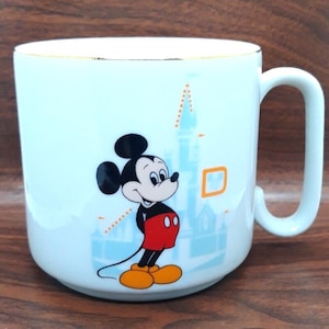 K&ouml;nnte beinhalten: Eine hellblaue Keramiktasse mit goldfarbenem Rand zeigt ein Cartoon-Bild von Mickey Mouse vor einer blauen und wei&szlig;en Illustration von Schloss Aschenputtel. Mickey Mouse tr&auml;gt seine charakteristischen roten Shorts und gelben Schuhe.