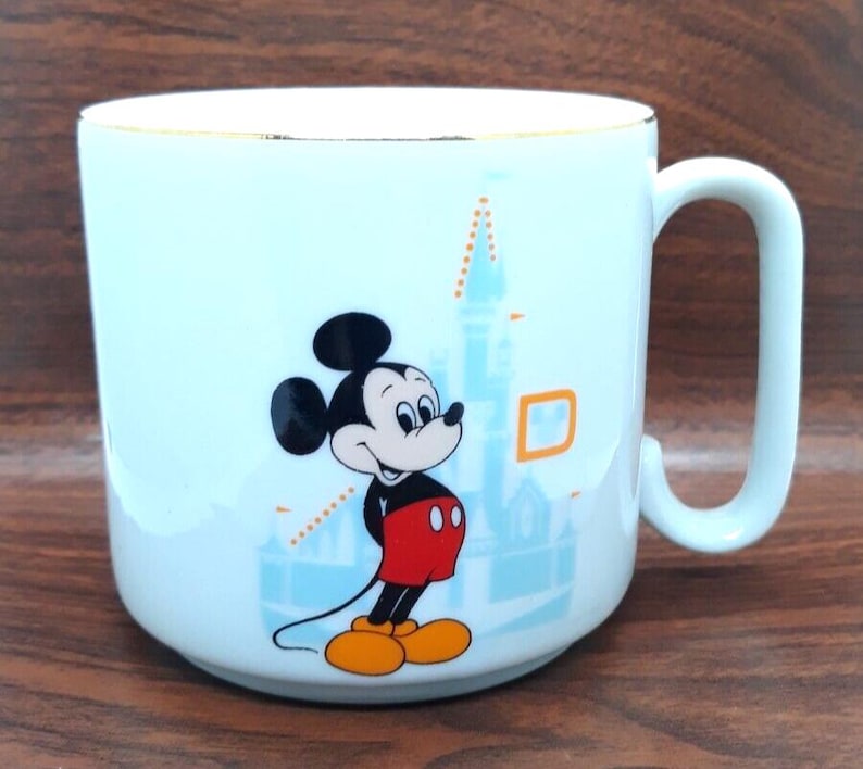 K&ouml;nnte beinhalten: Eine hellblaue Keramiktasse mit goldfarbenem Rand zeigt ein Cartoon-Bild von Mickey Mouse vor einer blauen und wei&szlig;en Illustration von Schloss Aschenputtel. Mickey Mouse tr&auml;gt seine charakteristischen roten Shorts und gelben Schuhe.