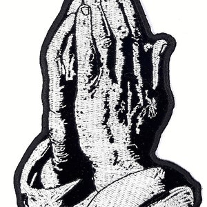 May include: Embroidered patch featuring a detailed depiction of hands clasped in prayer. The design is rendered in black and white, with intricate stitching creating a textured effect. The patch has a black border.