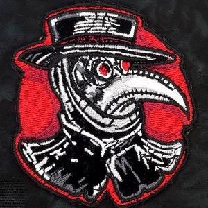 May include: Embroidered patch of a plague doctor wearing a black hat and a long white beak mask with red accents. The patch is circular and has a red background.