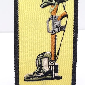 May include: A yellow embroidered patch with a black border depicting a soldier's boot, rifle, and helmet. The soldier is kneeling with their head bowed, representing a fallen soldier.