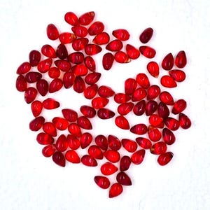 May include: A collection of small, teardrop-shaped, translucent red beads scattered on a white surface. The beads have a glossy finish and vary slightly in shade, ranging from deep crimson to lighter ruby tones. Ideal for jewelry making or crafting.