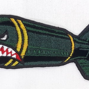 May include: Green embroidered patch of a cartoon bomb with a toothy, angry face. The bomb has yellow stripes and a black fuse.