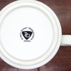 K&ouml;nnte beinhalten: Wei&szlig;e Keramiktasse mit einem schwarzen Walt Disney Productions Logo und dem Text "Japan" auf der Unterseite.