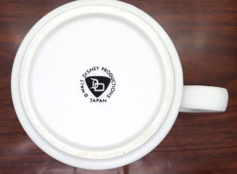 K&ouml;nnte beinhalten: Wei&szlig;e Keramiktasse mit einem schwarzen Walt Disney Productions Logo und dem Text "Japan" auf der Unterseite.