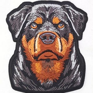 May include: Embroidered patch featuring a Rottweiler dog's face. The patch has a black border, with the dog's fur in shades of black, brown, and white. The dog's eyes are a light orange color, and the nose is brown.