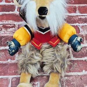 May include: A plush lion doll with a blue hat, white mane, and a red vest with gold trim. The doll has yellow arms and legs, and brown fur on the legs. The doll is set against a red brick wall.
