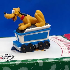 May include: A Pluto figurine, part of a "Merry Miniatures" collection, rests on a blue and white train car. The yellow dog figure has a red collar and black eyes. The train car has black wheels and is on a green box.