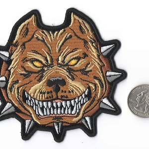 May include: Embroidered patch featuring a snarling dog's head with sharp teeth and yellow eyes. The patch is brown with a black border and silver spikes around the neck. A U.S. quarter is next to the patch for size comparison.