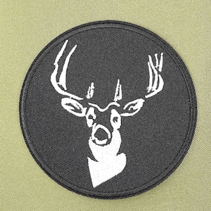 May include: A circular black patch featuring a white deer head with large antlers. The patch has a black border and is set against a light olive green background. The deer's head is detailed with white lines.
