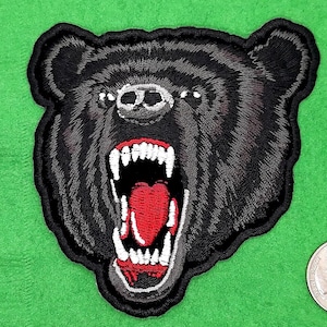 May include: Embroidered patch featuring a snarling black bear with a wide-open mouth, revealing white teeth and a red interior. The bear's fur is detailed with gray shading, and the patch has a black border. The patch is on a green background.