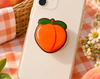 May include: A clear smartphone case featuring a circular peach-shaped grip attachment on the back. The peach graphic is orange with a dark brown outline and two green leaves at the top, centered on the back of the mobile device.