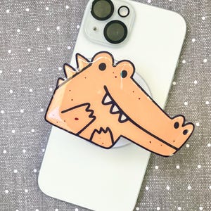 May include: A light orange alligator phone grip attached to a white smartphone. The alligator has black outlines and a cartoonish design. The phone is on a gray dotted surface.
