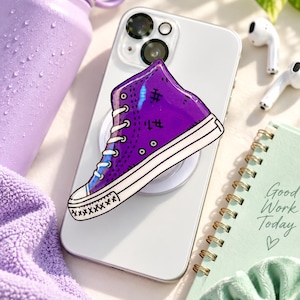 May include: A purple high-top sneaker phone grip with white laces and a white rubber sole, featuring black stitched details, attached to the back of a white smartphone. The sneaker design has a glossy finish and a distinct cartoon-like aesthetic.