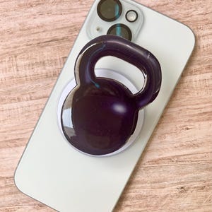 May include: A white smartphone with a black and purple kettlebell phone grip attached. The kettlebell grip is round with a handle and is centered on the phone. The phone is on a light brown wooden surface.