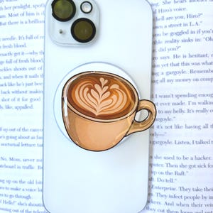 May include: A white smartphone case featuring a circular phone grip attachment. The grip displays a graphic illustration of a tan ceramic coffee cup filled with a latte, showcasing intricate white foam art in the shape of a leaf pattern.
