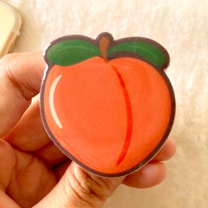 May include: A peach-shaped sticker with a vibrant orange body, a green leaf crown, and a brown stem. The sticker has a white border and a vertical red line down the center. The sticker is held in a hand.