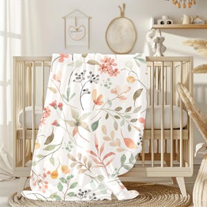 May include: A white baby blanket with a watercolor floral pattern featuring pink, orange, and green flowers and leaves.