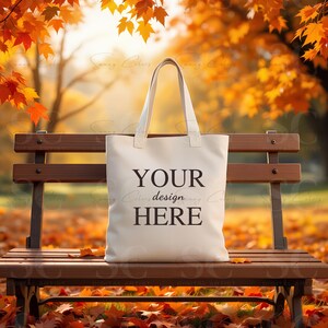 May include: Cream-colored tote bag with the text "YOUR design HERE" printed in black. The bag has two handles and is sitting on a wooden bench in a park with autumn leaves.