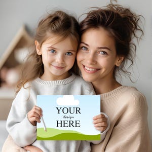 May include: A rectangular sign featuring a light blue sky with a white cloud above rolling green hills, with the black text "YOUR design HERE" centred in the middle. The sign is held by two individuals, one wearing a white jumper and one in beige.