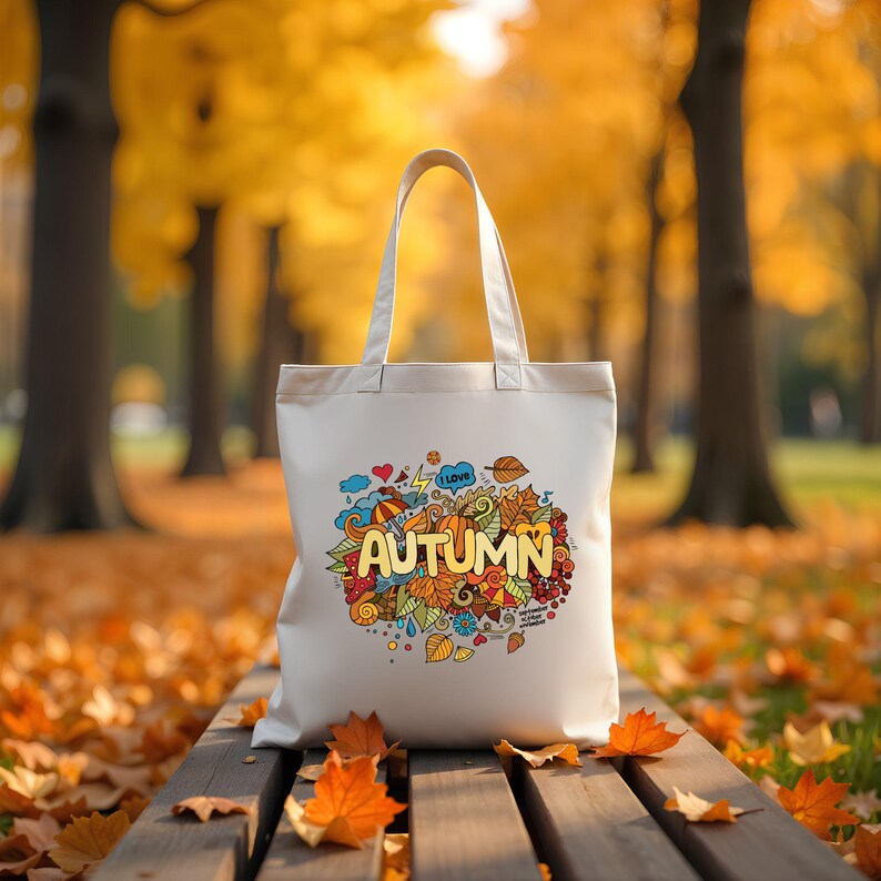May include: A cream-colored tote bag with a colorful autumn-themed design. The design includes the word "AUTUMN" in yellow, surrounded by leaves, an umbrella, and other fall-related elements. The bag is sitting on a wooden bench.