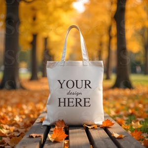 May include: A cream-colored tote bag with the text "YOUR design HERE" printed on the front. The bag has two handles and is sitting on a wooden bench covered in fallen autumn leaves. The background features a blurred view of trees with yellow foliage.
